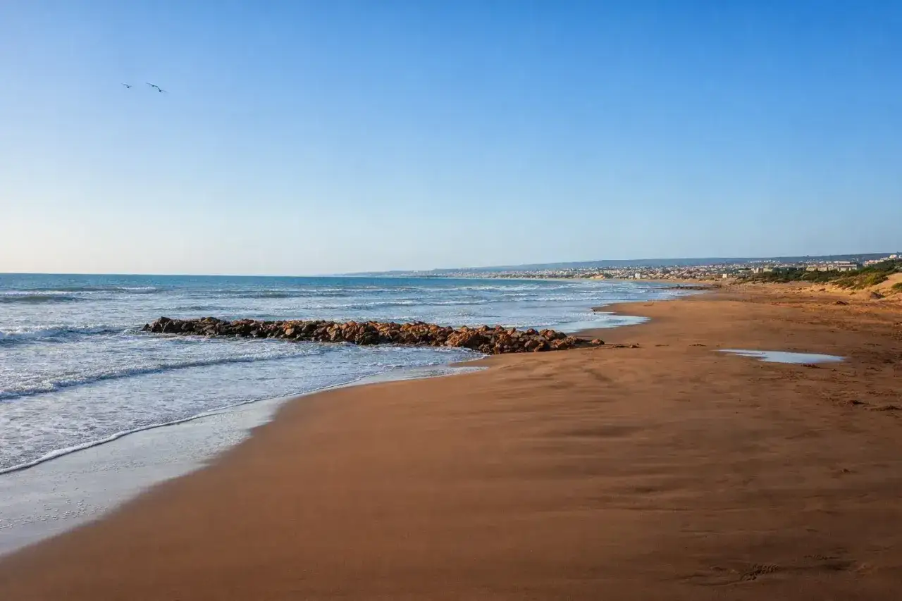 Spiagge bianche di Ragusa bagnate da un mare azzurro, con un molo di sassi e gabbiani in volo.