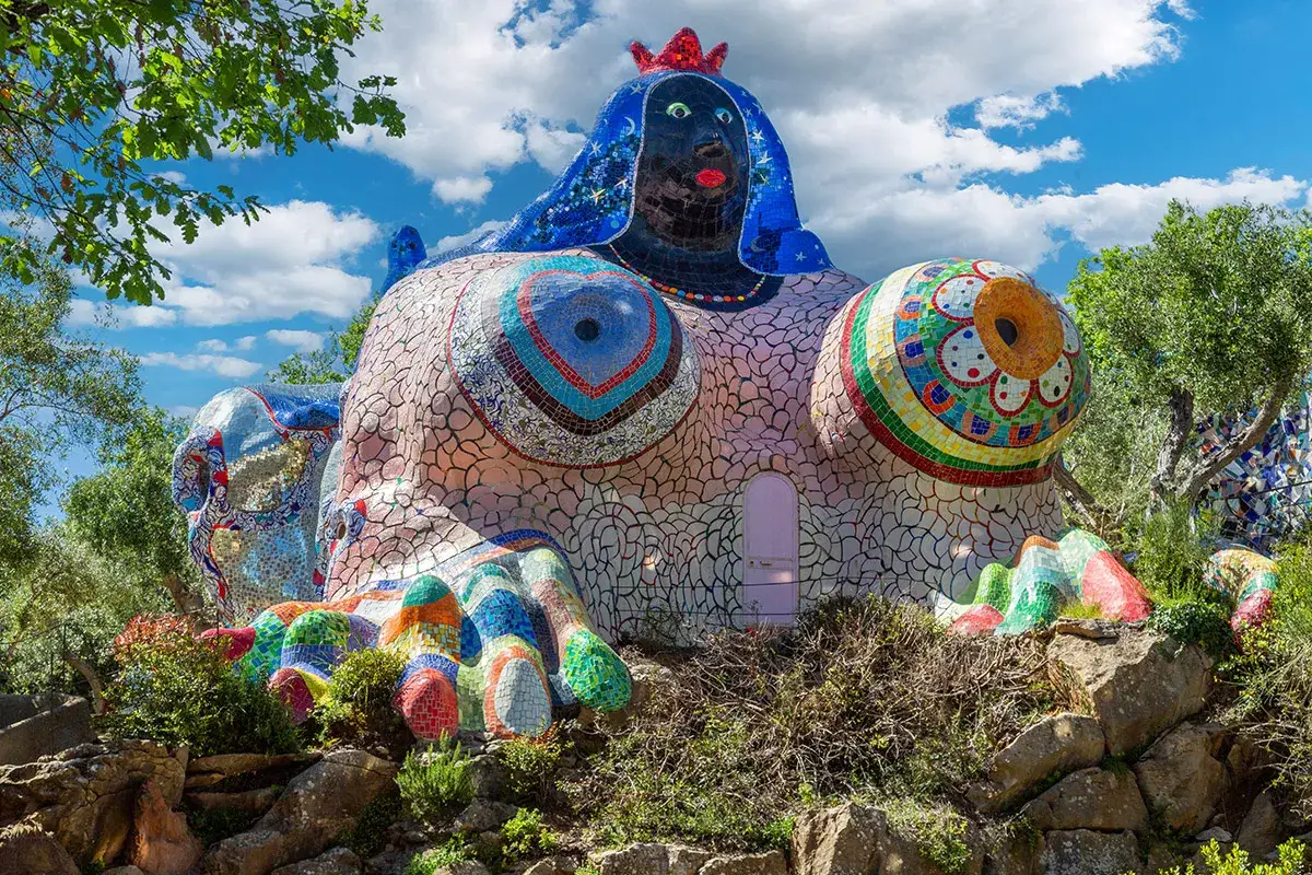 La figura colorata e sinuosa del Giardino dei Tarocchi in Toscana, con la sua testa coronata e i seni decorati, emerge tra la vegetazione.
