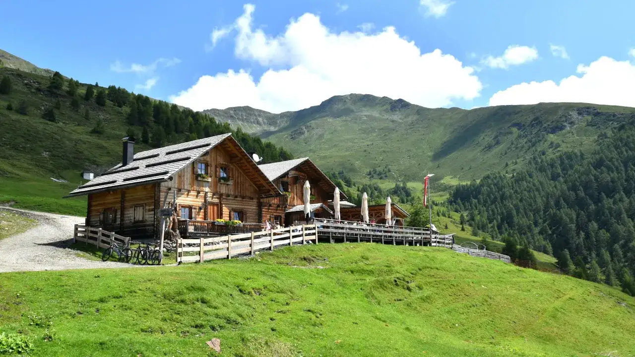 Rifugio in legno con tavoli all'aperto, ideale per escursioni in Alto Adige. Montagne verdi e cielo azzurro.