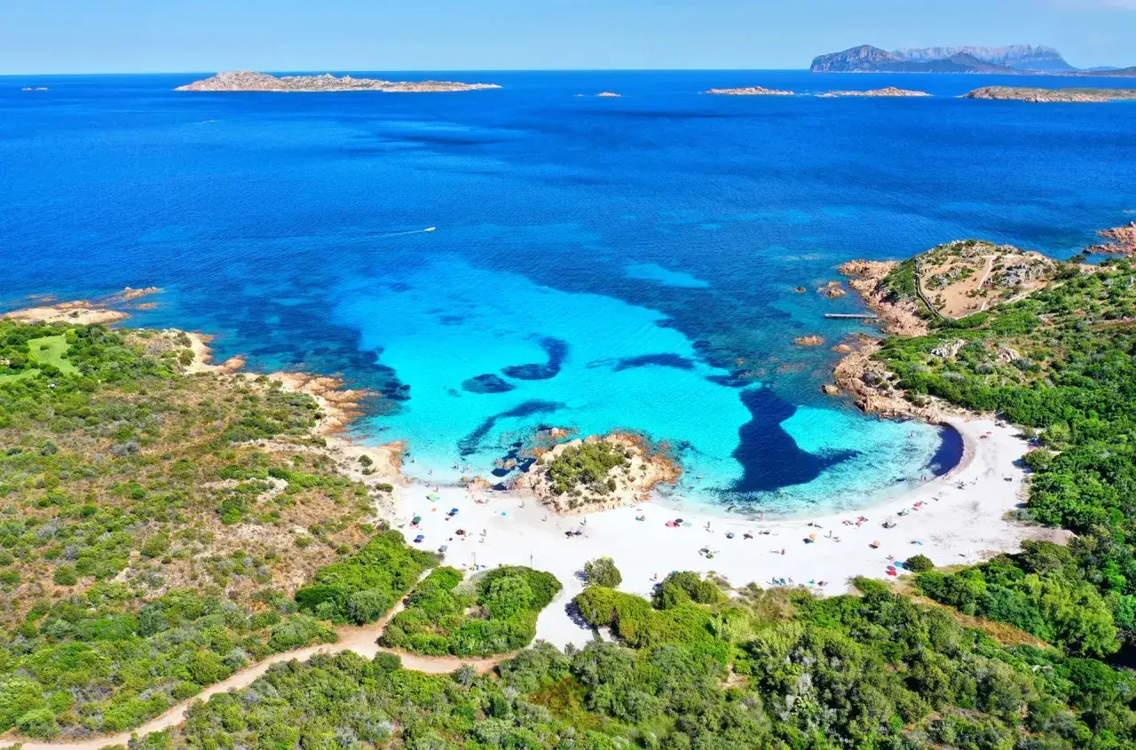 Vista aerea di una baia con sabbia bianca e acque turchesi cristalline, circondata da macchia mediterranea. Un vero gioiello tra le spiagge più belle della Sardegna.