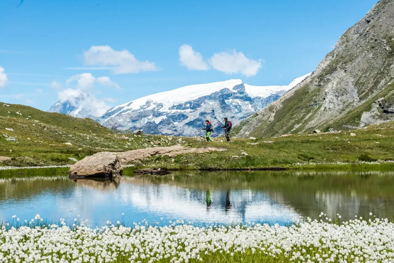 Escursionisti nel Val d'Ayas, con laghetto alpino e montagne innevate sullo sfondo.