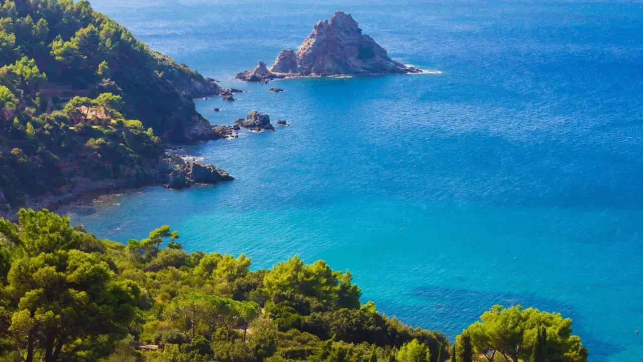 Scogliera verdeggiante della Maremma Toscana si tuffa nel mare azzurro, con un'isola rocciosa che emerge dalle acque limpide.