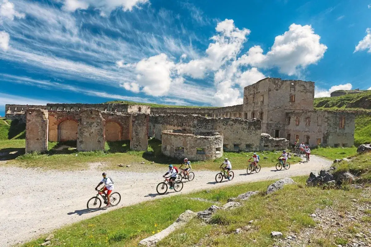 Ciclisti esplorano il via del sale cammino tra le rovine di un antico forte, sotto un cielo azzurro con nuvole bianche.