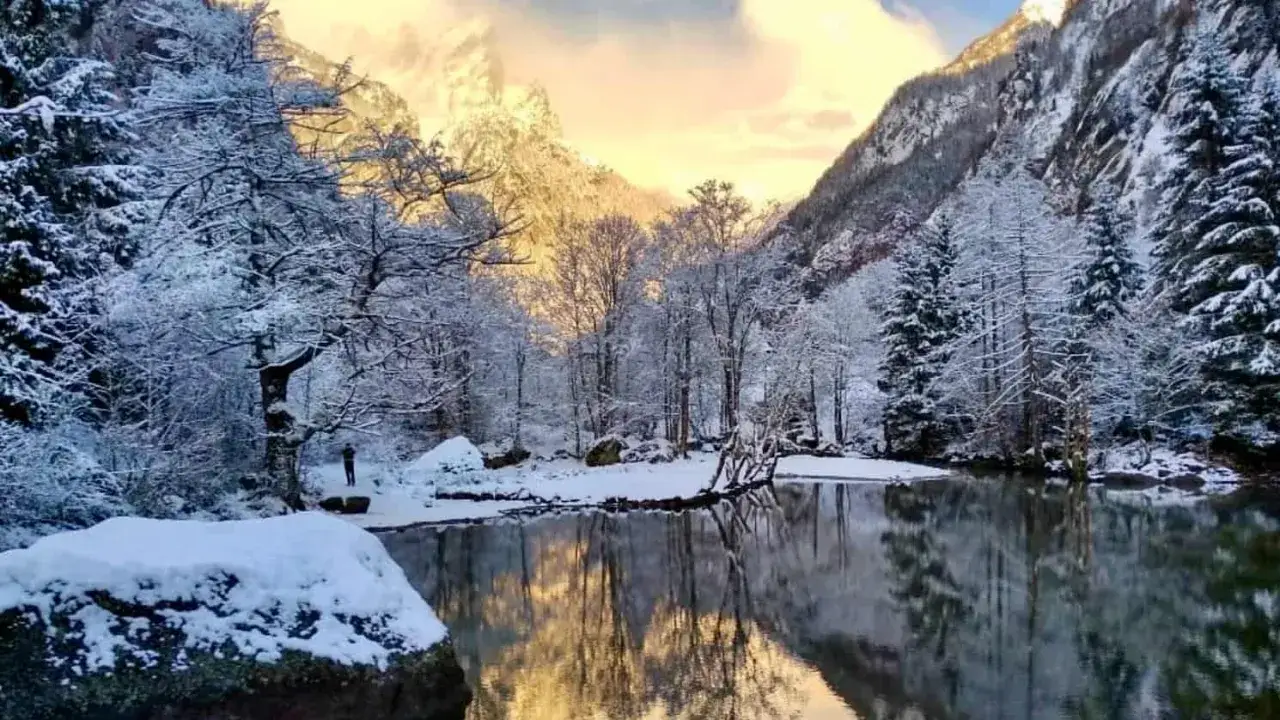 Un solitario escursionista ammira il magico paesaggio del Val di Mello inverno, con alberi innevati che si specchiano in un lago tranquillo sotto un cielo dorato.