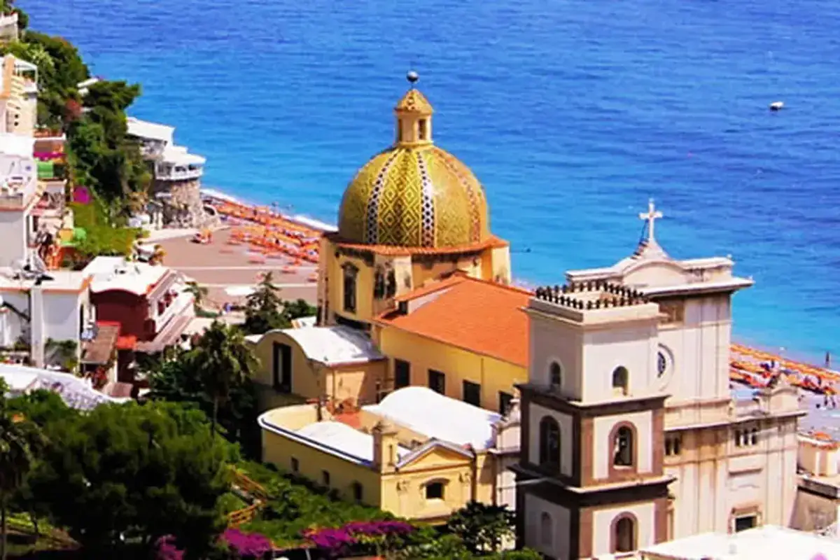 Cosa vedere a Positano: la cupola maiolicata della chiesa di Santa Maria Assunta domina il panorama, con la spiaggia e il mare blu sullo sfondo.
