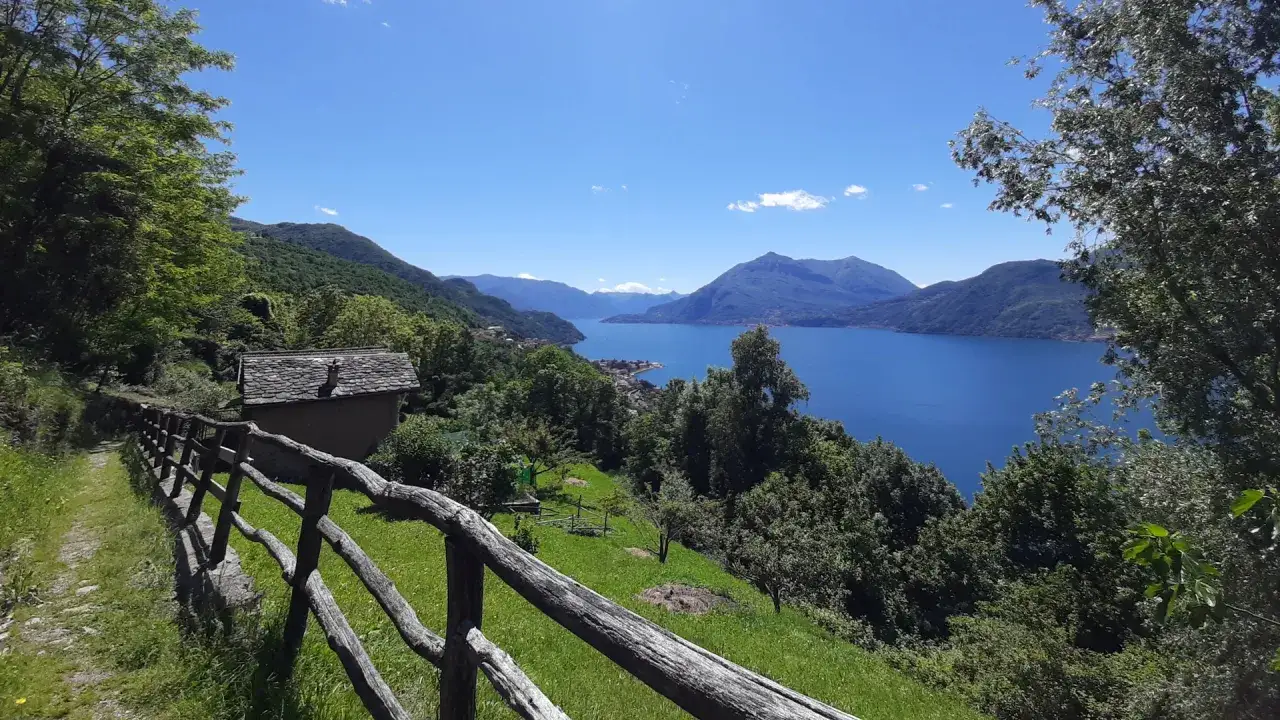 Il sentiero del viandante serpeggia tra il verde, con una casetta in pietra e un lago azzurro sullo sfondo.