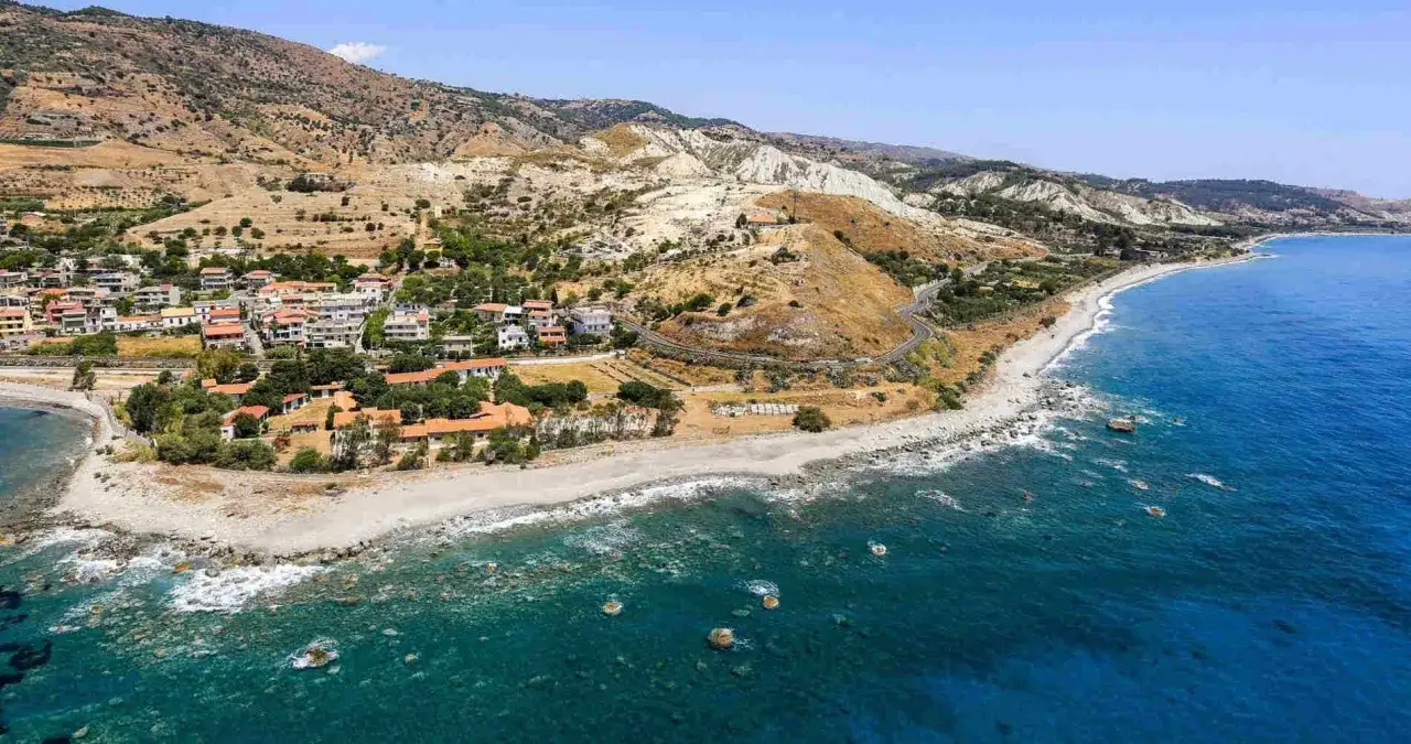 Panoramica della Calabria Costa Ionica: un borgo aggrappato alla collina digrada verso il mare blu cobalto, con una spiaggia di ciottoli e rocce.
