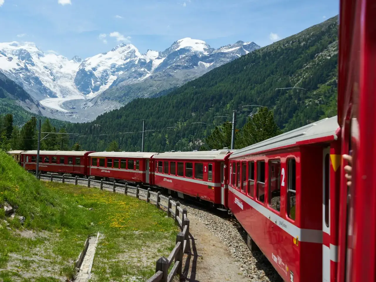 Un treno rosso attraversa un paesaggio alpino con montagne innevate e foreste verdi.