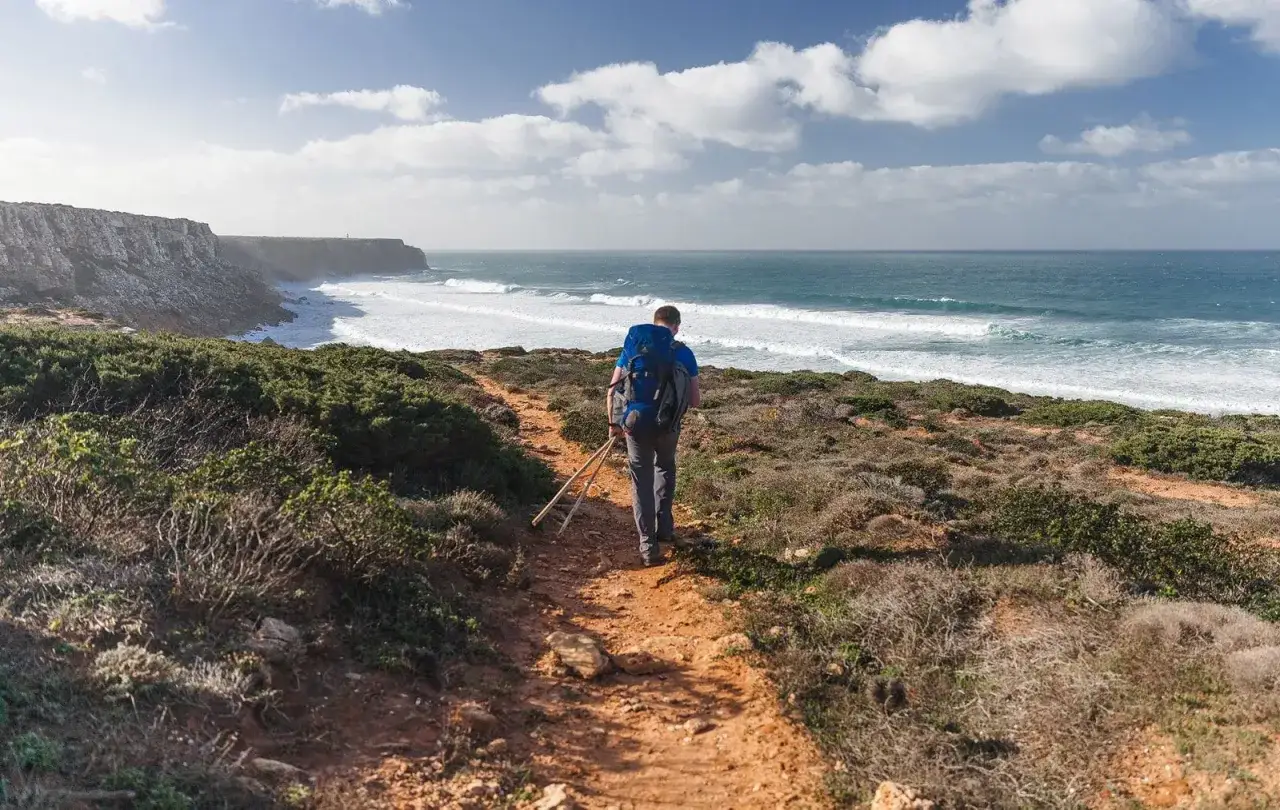 Escursionista con zaino e bastoncini lungo un sentiero costiero, godendosi le onde dell'oceano. Le tappe della Rota Vicentina offrono panorami mozzafiato.