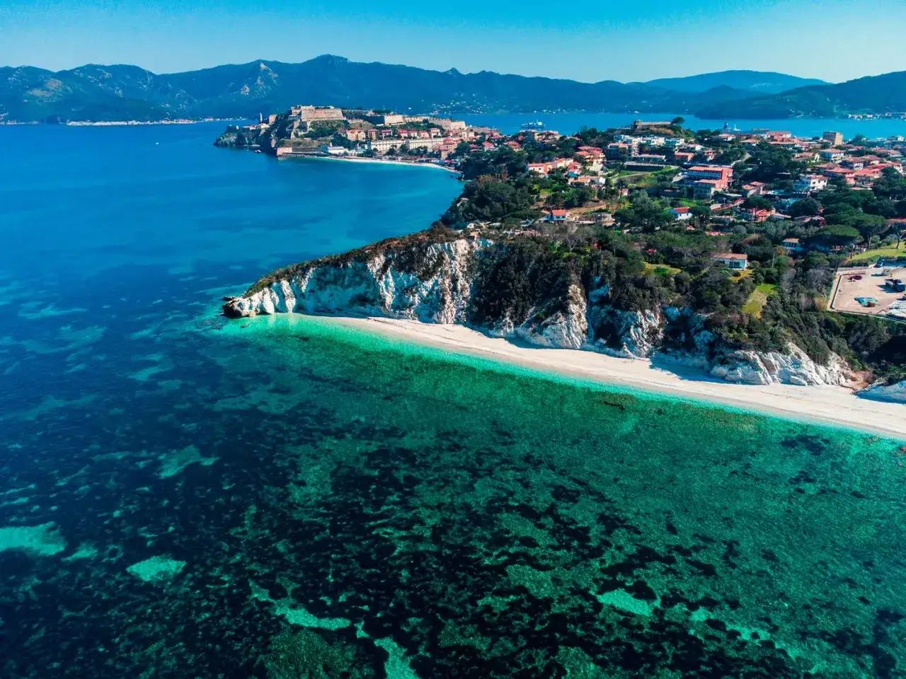 Vista aerea di una spiaggia bianca su un'isola vicino all'Elba, con acque cristalline e un borgo arroccato su una collina.