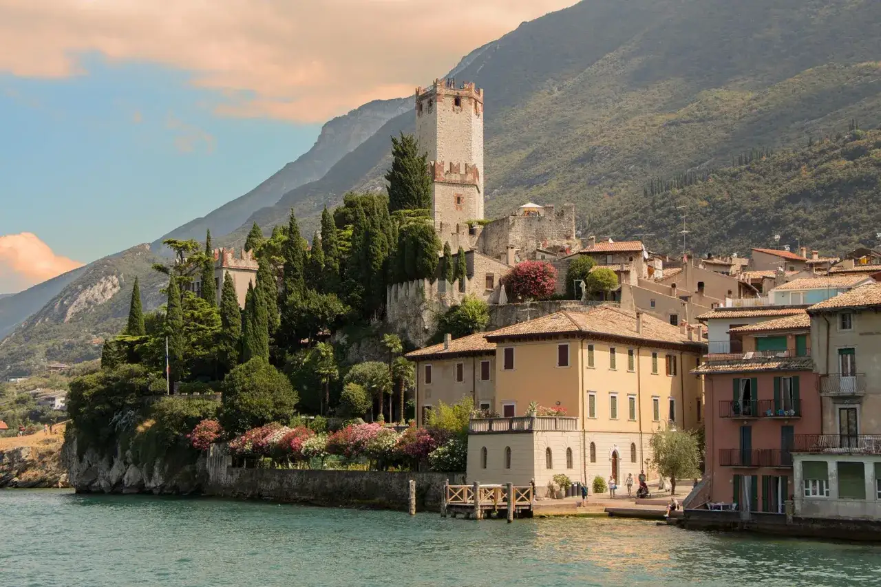 Borghi da visitare nel Nord Italia: un castello medievale domina un pittoresco paese affacciato sul lago, tra cipressi e fiori colorati.