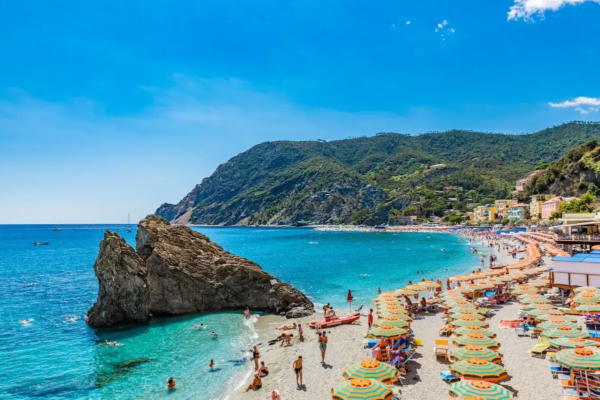 Spiaggia affollata sulla riviera di Levante, con ombrelloni colorati, mare azzurro e colline verdi sullo sfondo.