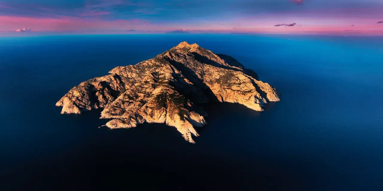 L'isola di Montecristo, un gioiello roccioso nel blu intenso del mare, si staglia sotto un cielo rosa e arancio al tramonto.