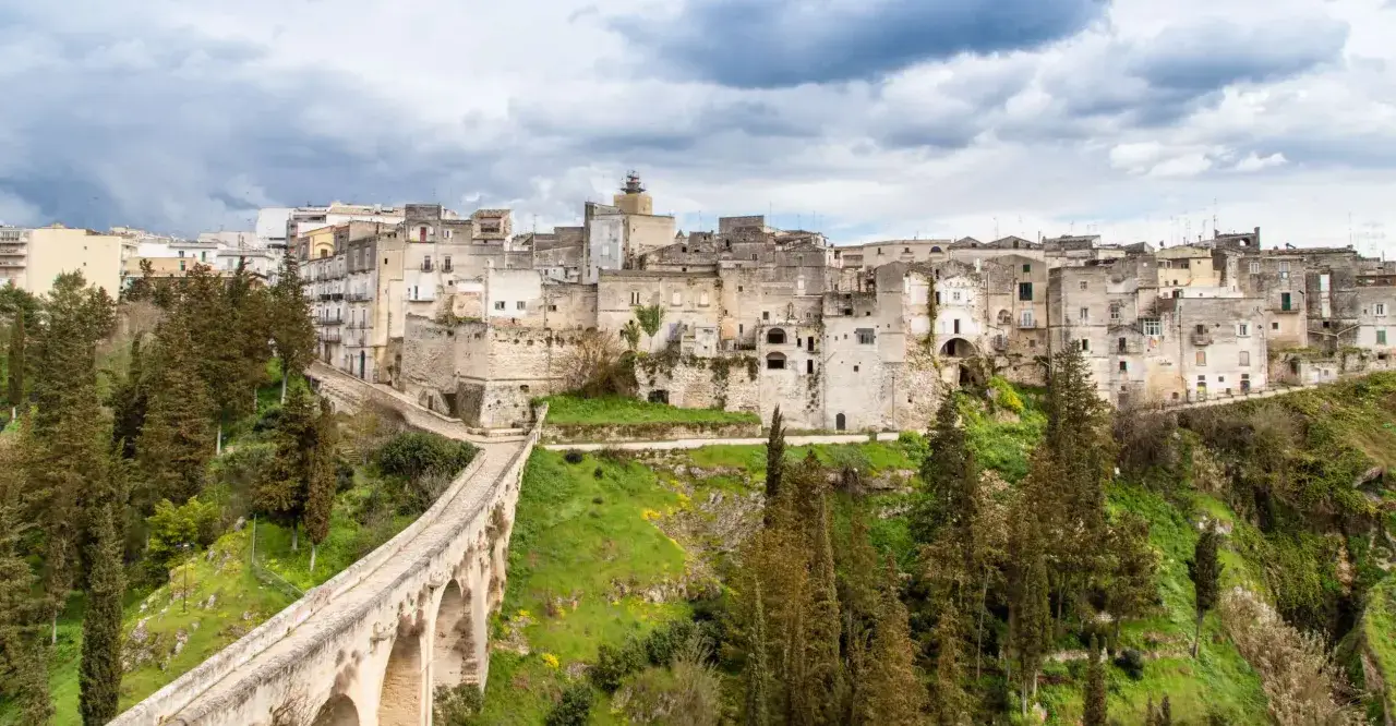 Ponte acquedotto e case scavate nella roccia nel centro storico di Gravina in Puglia, con alberi e cielo nuvoloso.