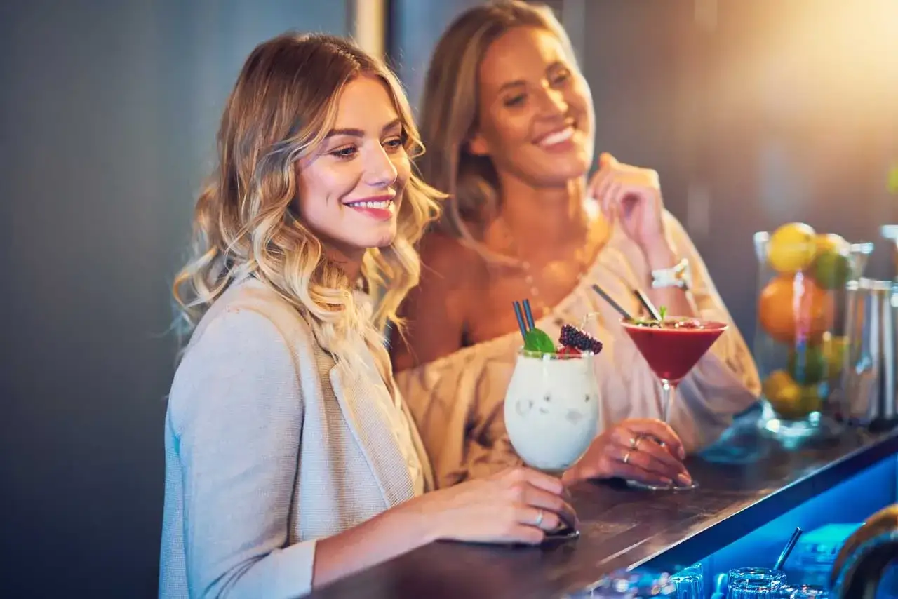 Due amiche sorridono al bancone di un locale, gustando cocktail colorati. Un assaggio della vivace firenze vita notturna.