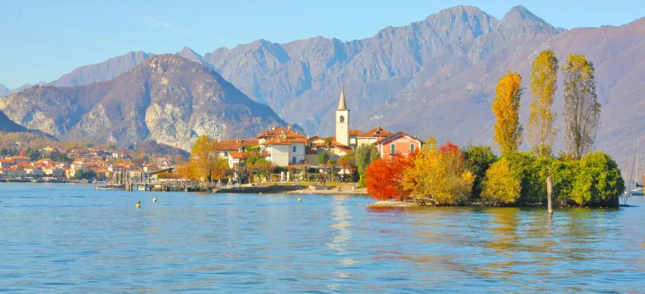 Island village with colorful autumn trees on a lake, with mountains in the background.