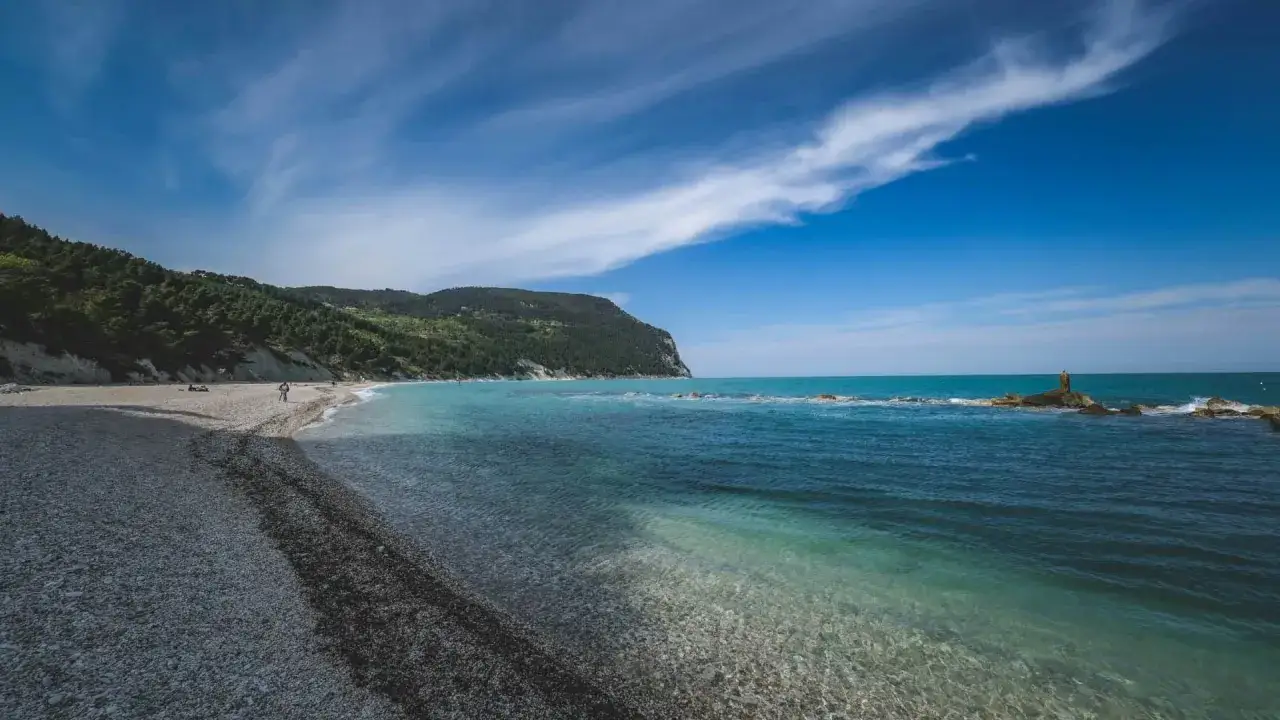 Spiaggia di ciottoli e mare cristallino, un luogo perfetto per scoprire cosa fare nelle Marche, con una scogliera verdeggiante sullo sfondo.