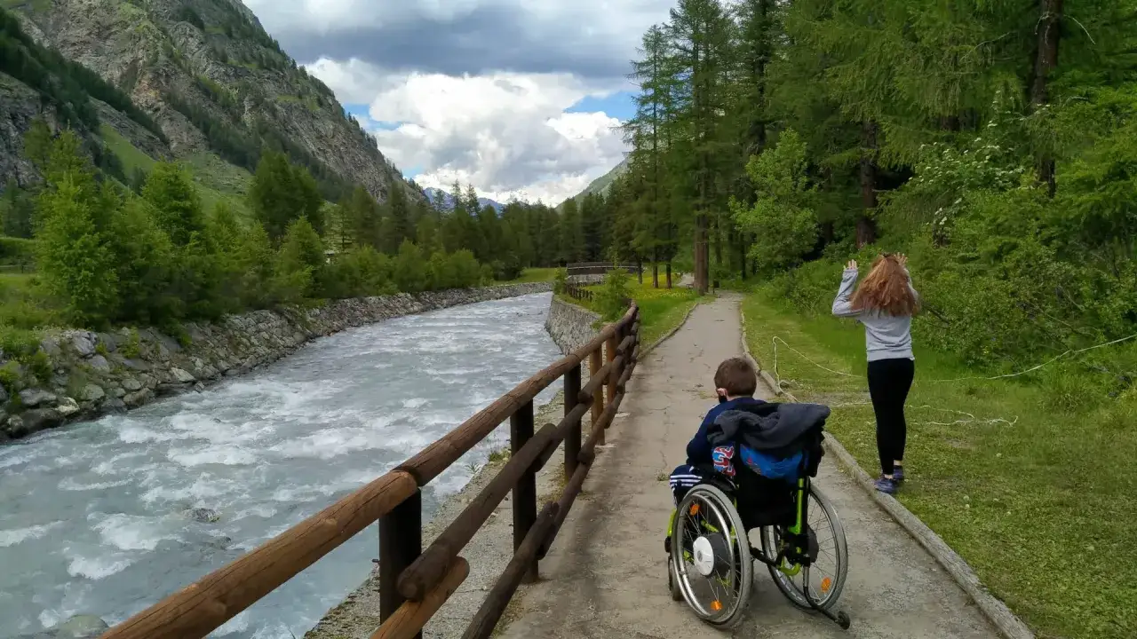 Un bambino in carrozzina e una ragazza camminano lungo un sentiero nel Val di Rhemes, con un fiume impetuoso e montagne verdi sullo sfondo.