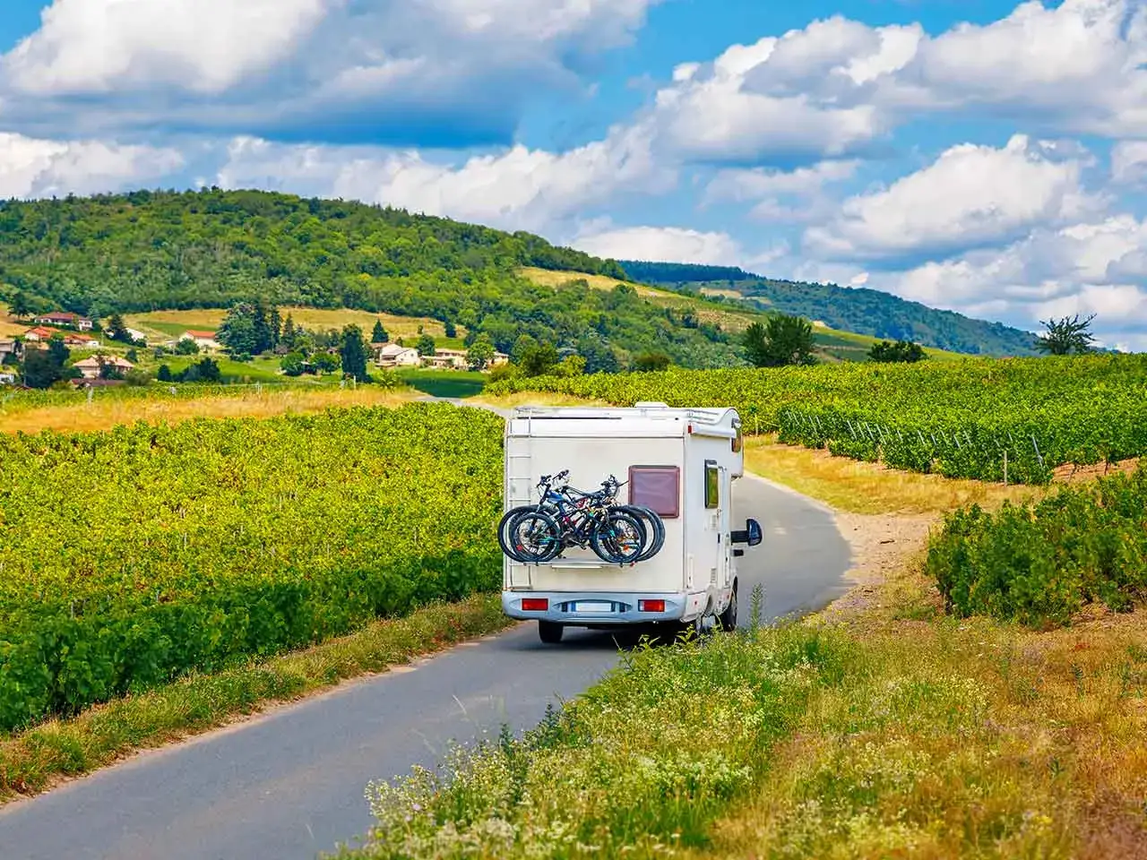 Un camper con biciclette sul retro percorre una strada tra rigogliosi vigneti, con colline verdi e un cielo azzurro punteggiato di nuvole. Un'avventura in Toscana in camper.