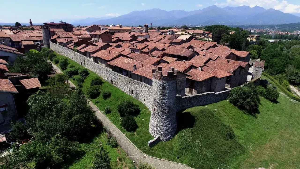 Antica cinta muraria con torre circolare, parte del ricetto di Candelo, si staglia contro un cielo azzurro e montagne sullo sfondo.