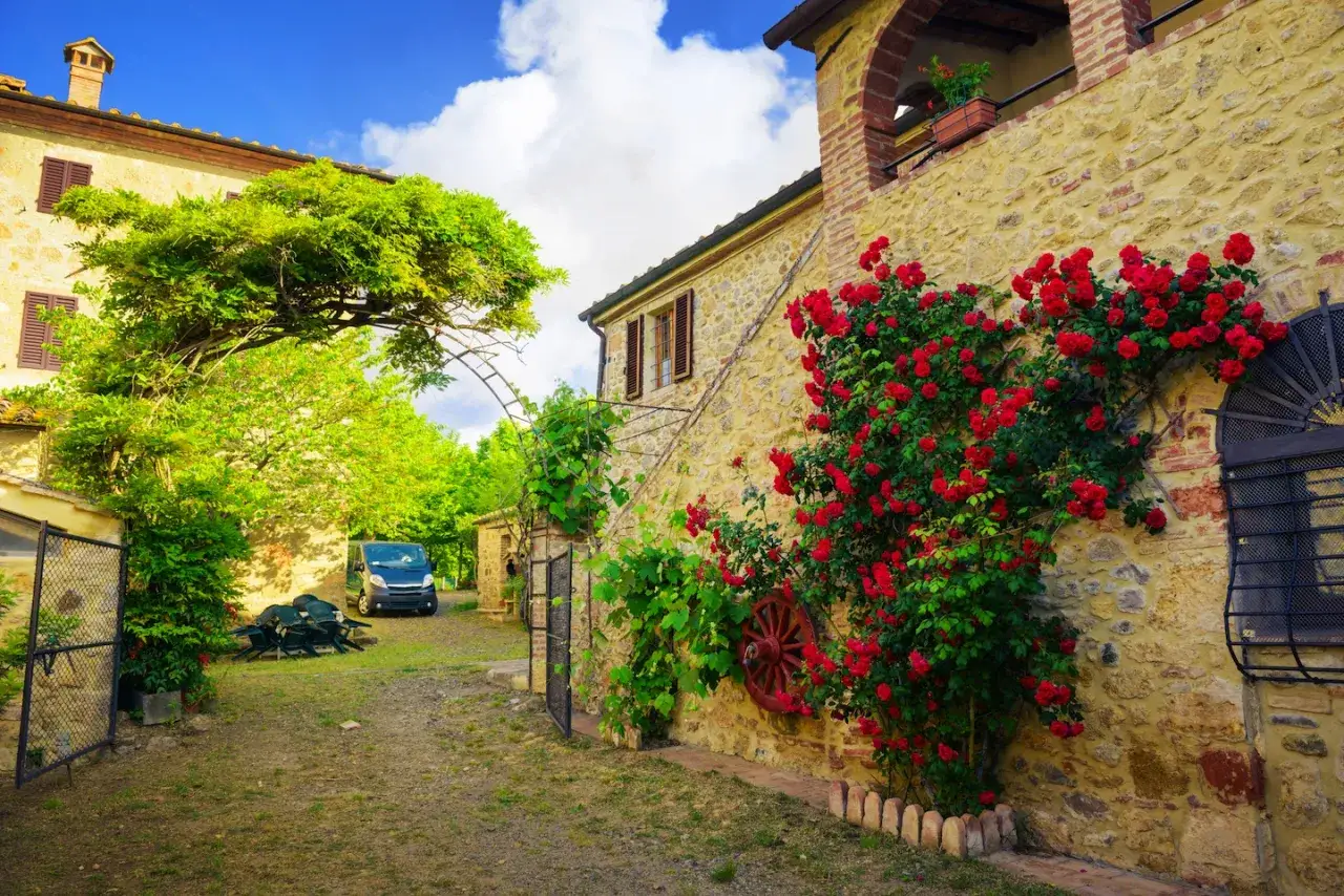 Un casolare in pietra con rose rosse rampicanti, un'auto parcheggiata e un cielo azzurro: i sogni del Chianti prendono vita.