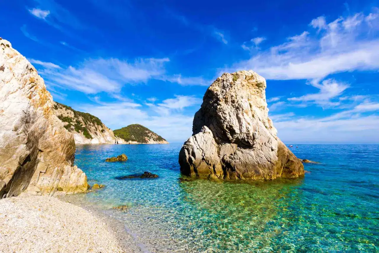 Scogli imponenti e acque cristalline nel golfo di Follonica, con colline verdi sullo sfondo sotto un cielo azzurro e nuvoloso.