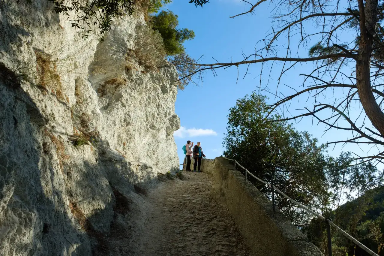 Due escursionisti percorrono un antico sentiero del pellegrino, fiancheggiato da una parete rocciosa imponente e alberi spogli sotto un cielo azzurro.