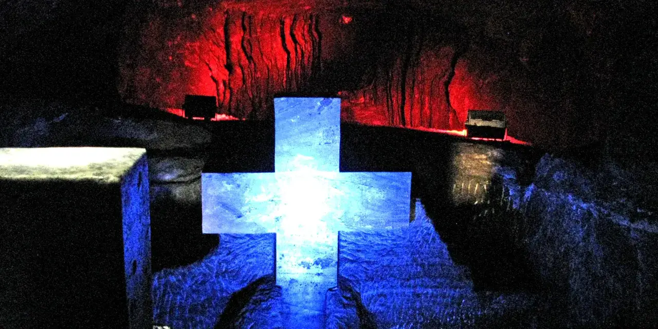 Croce luminosa blu al centro, con pareti rosse e nere illuminate in profondità. Un'atmosfera mistica nella cattedrale del sale.