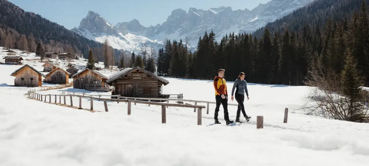 Coppia in cammino tra baite innevate e montagne maestose, un'esperienza di escursioni invernali in Trentino.