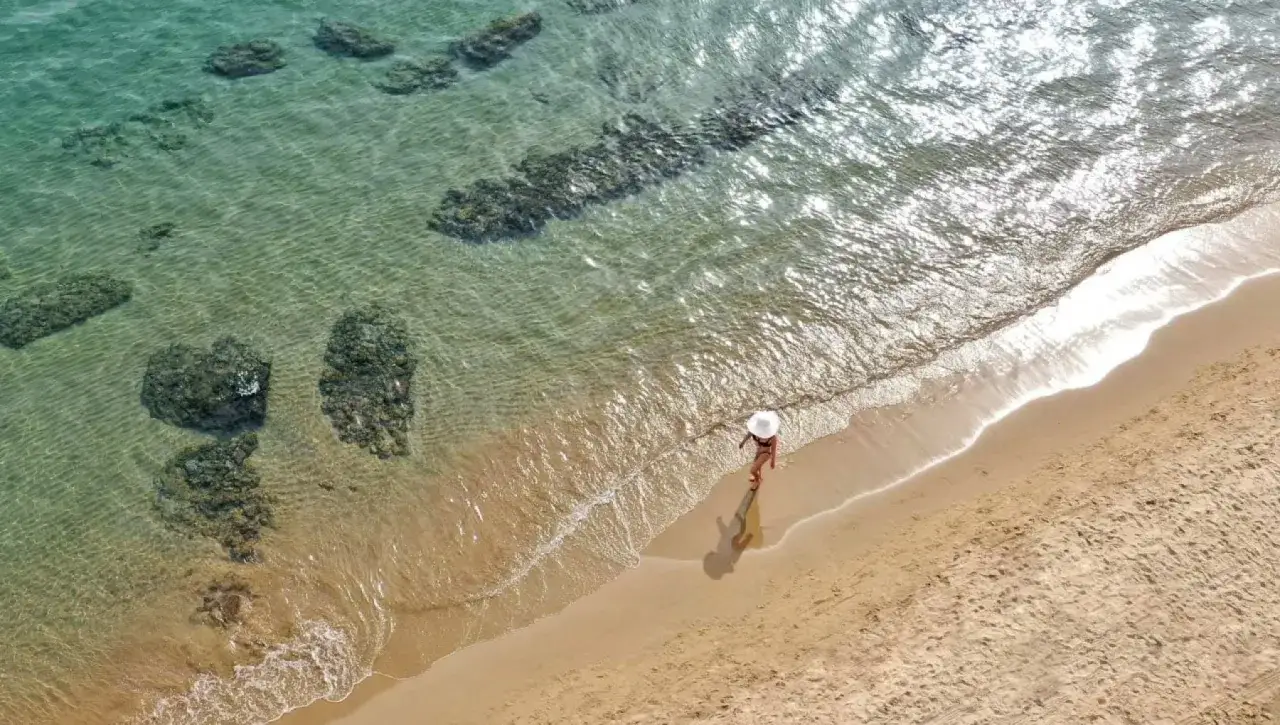Una donna con cappello bianco cammina sulla sabbia dorata, bagnata dalle acque cristalline di un mare bello nel Lazio. Rocce affiorano dall'acqua trasparente.