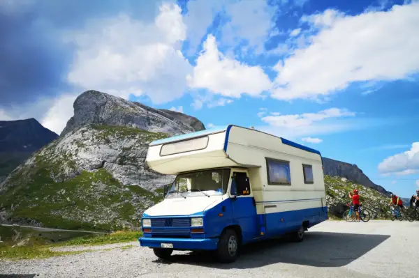Camper blu e bianco parcheggiato in montagna, pronto per esplorare gli **itinerari camper Italia**. Due ciclisti sullo sfondo.