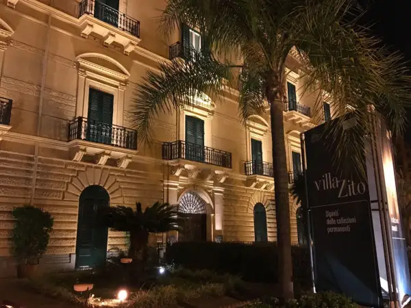 Villa Zito a Palermo, un'elegante dimora illuminata di notte, con palme che incorniciano la facciata e un cartello che annuncia "Dipinti delle collezioni permanenti".