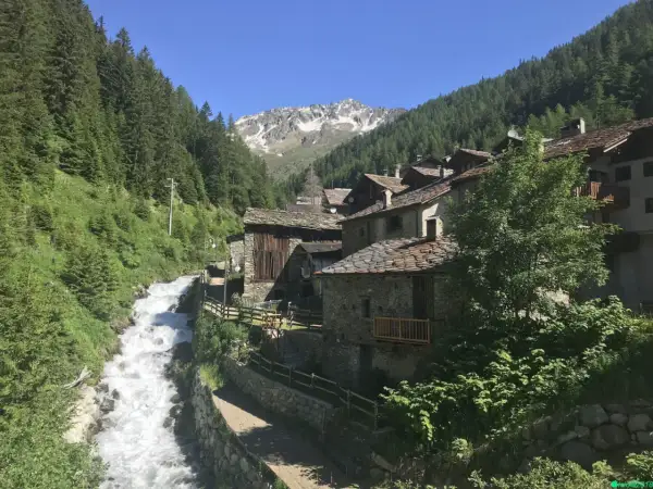 Un pittoresco borgo alpino nel Nord Italia, con case in pietra e tetti in ardesia, incastonato tra boschi rigogliosi e un torrente impetuoso.
