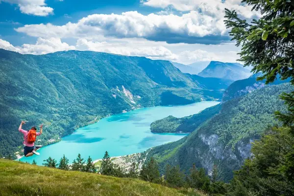 Giovane salta felice con zaino rosso, godendosi le passeggiate facili vicino a Trento con vista su lago turchese e montagne verdi.