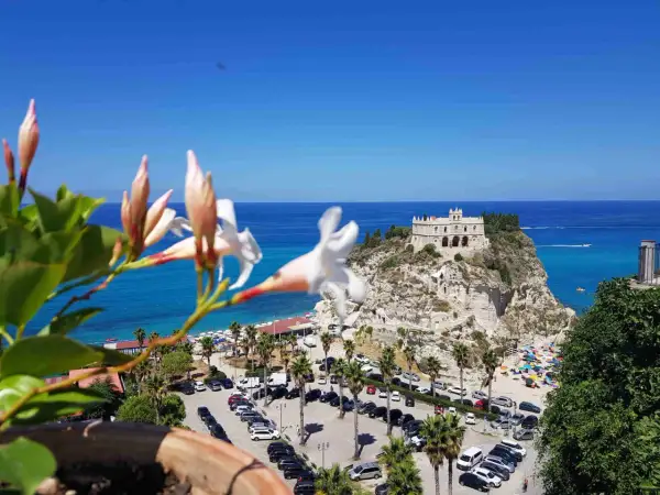 Fiori bianchi in primo piano, sullo sfondo il Santuario di Santa Maria dell'Isola a Tropea, un must per chi cerca cosa vedere in Calabria. Spiaggia affollata e mare azzurro.