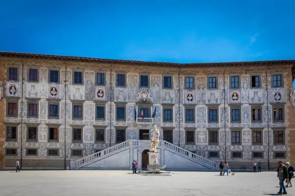 Palazzo della Sapienza a Pisa, con la sua facciata decorata, si affaccia su una piazza dove si intravede la **torre di Pisa** in lontananza, nella **Piazza del Duomo, Pisa, PI**.