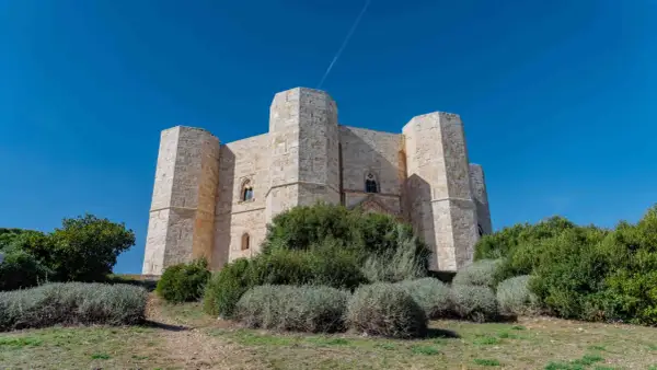 Castel del Monte, un'imponente fortezza ottagonale in pietra chiara, si erge maestoso sotto un cielo azzurro intenso, circondato da vegetazione mediterranea.