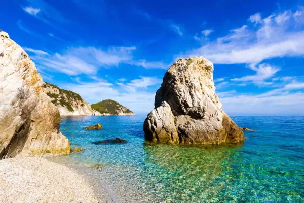 Scogli imponenti e un mare cristallino toscana, con acque turchesi che lambiscono una spiaggia di ciottoli bianchi sotto un cielo azzurro.