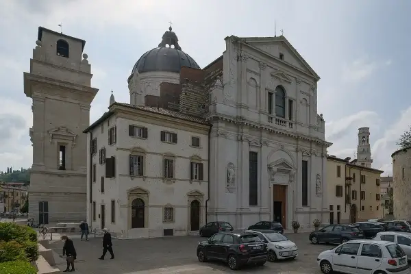La piazza di San Giorgio a Verona, con la chiesa imponente, la torre campanaria e alcune persone che passeggiano tra le auto parcheggiate.