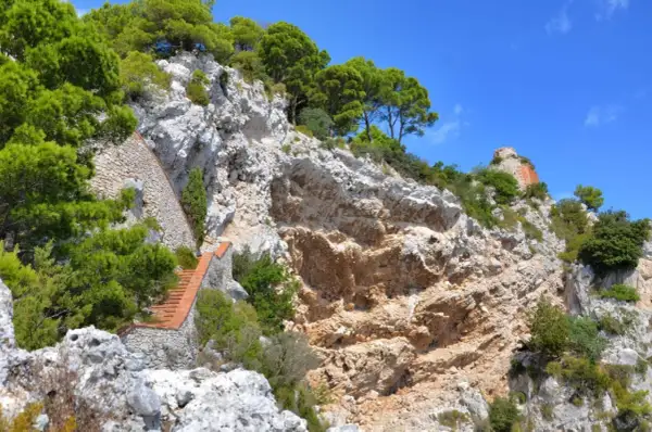 Scogliera rocciosa con una scalinata in mattoni che sale verso la cima, dove si intravede un rudere. Si immagina il **salto di Tiberio**.