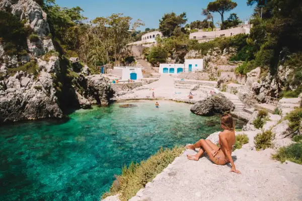 Una donna ammira le acque cristalline di una baia pugliese, con piccole spiagge sabbiose e case bianche arroccate sulla scogliera.