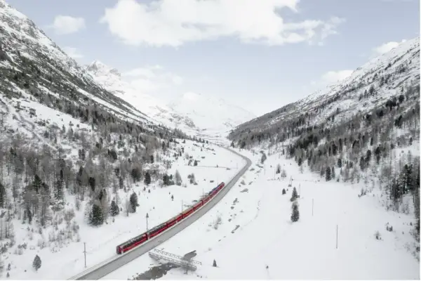 Un treno rosso attraversa una valle innevata, un viaggio tra i **treni panoramici Italia** con montagne maestose e alberi spogli.