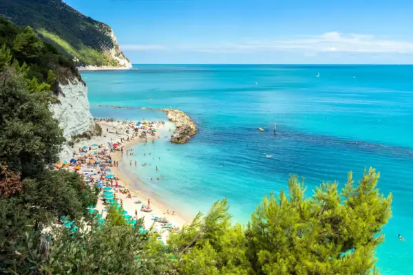 Spiaggia affollata con ombrelloni colorati e gente che prende il sole, bagnata da un **abruzzo mare bello** e cristallino, con scogliere bianche e vegetazione rigogliosa.