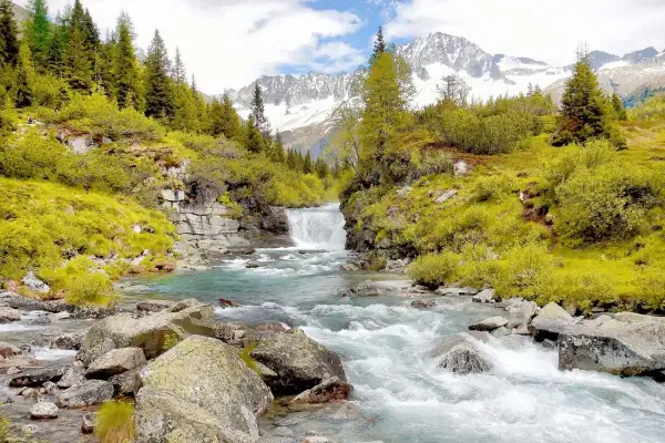 Cascata alpina e torrente cristallino, perfetti per gite in Trentino in giornata. Montagne innevate sullo sfondo.