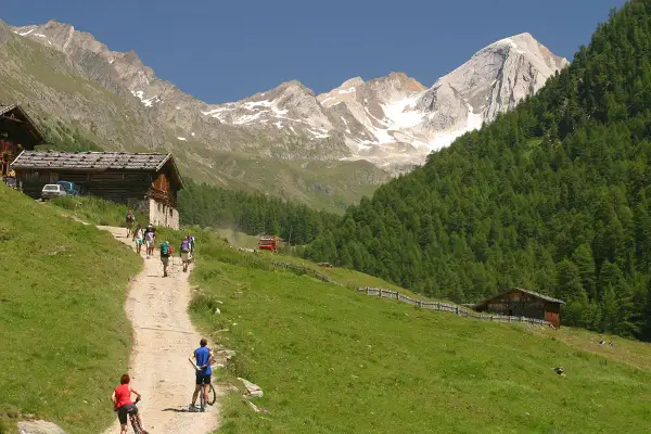 Escursionisti e ciclisti percorrono un sentiero nel **Val di Fosse**, tra baite in legno e montagne innevate.