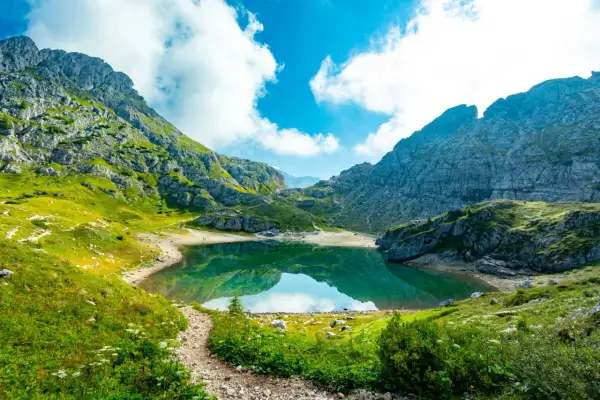 Sentiero in montagna tra verdi prati e rocce, con un lago alpino che riflette il cielo azzurro e nuvole bianche. Splendidi paesaggi naturali.