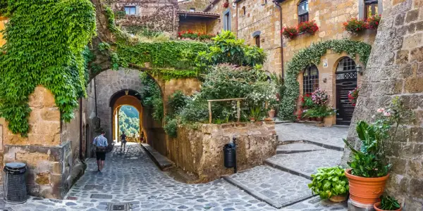 Un vicolo acciottolato in un borgo da visitare, con case in pietra ricoperte di edera e fiori. Un arco conduce a una vista panoramica.