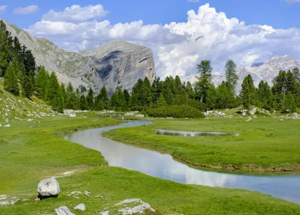 Un sereno sentiero di montagna serpeggia tra prati verdi e un ruscello cristallino, con imponenti vette sullo sfondo.