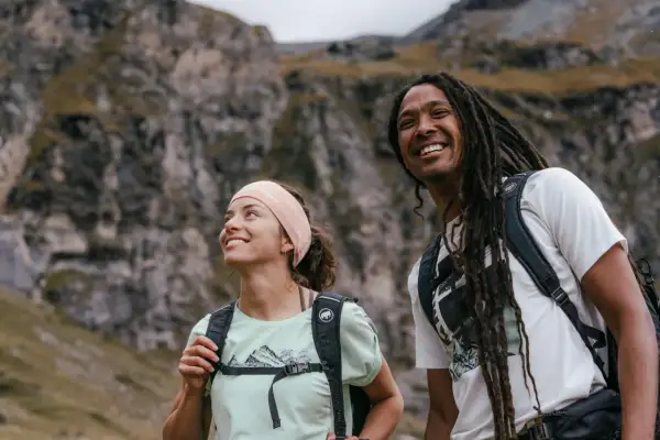Coppia sorridente in montagna, con zaini e abbigliamento montagna estate perfetto per l'avventura.