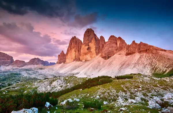 Le maestose cime di Lavaredo illuminate dal tramonto, con un cielo viola e blu che fa da sfondo a un paesaggio alpino mozzafiato.